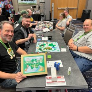 David and two players sit around a table with the board game Luck: Legends of Ireland set up in front of them. The board game has a path shaped like a clover and David is holding the cover of the game up. There are gamers playing other games in the background.
